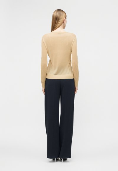 Γυναίκα με μακριά ξανθιά μαλλιά, που φοράει μπεζ μακρυμάνικη μπλούζα και φαρδιές μαύρες παντελόνες, στέκεται με την πλάτη προς τα εμπρός σε ένα απλό φόντο.