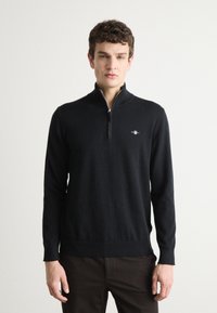 Crni džemper na patentni zatvarač od mekanog materijala, s visokim ovratnikom, dugim rukavima te rebrastim rubovima na manžetama i donjem dijelu. Mali logo na prsima.