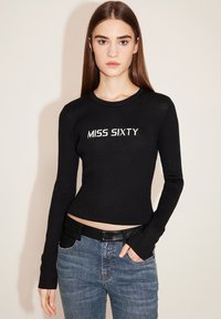 Schwarzes, geripptes Langarmoberteil mit dem aufgestickten Schriftzug "MISS SIXTY" in Weiß, kombiniert mit blauen Jeans und einem schwarzen Gürtel.