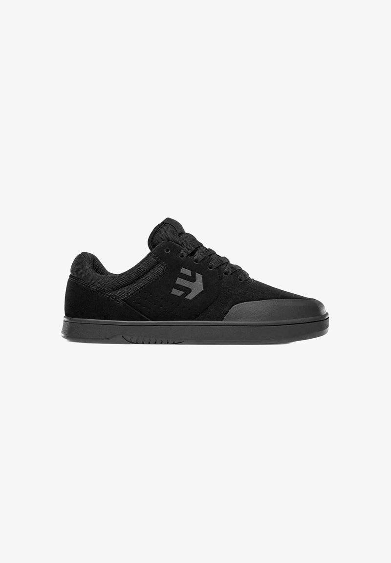 Zapatilla baja negra de materiales de ante y malla, con cordones en la parte delantera, suela de goma y un logotipo estilizado en el lateral.