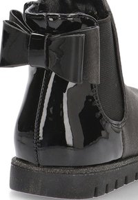 Bota de charol negro con parte superior de tejido texturizado y un gran lazo en la parte trasera. Presenta una suela de goma plana y un acabado liso.