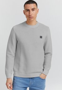 Maglione grigio chiaro con texture a coste, scollo a girocollo e una piccola etichetta nera sul petto. Abbinato a pantaloni in denim blu.