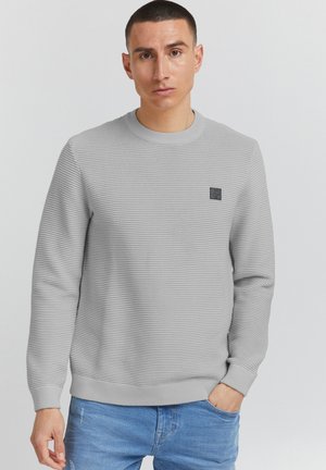 Lichtgrijze trui met een geribbelde structuur, ronde hals en een klein zwart logo-opdruk op de borst. Gepaard met blauwe denim broek.