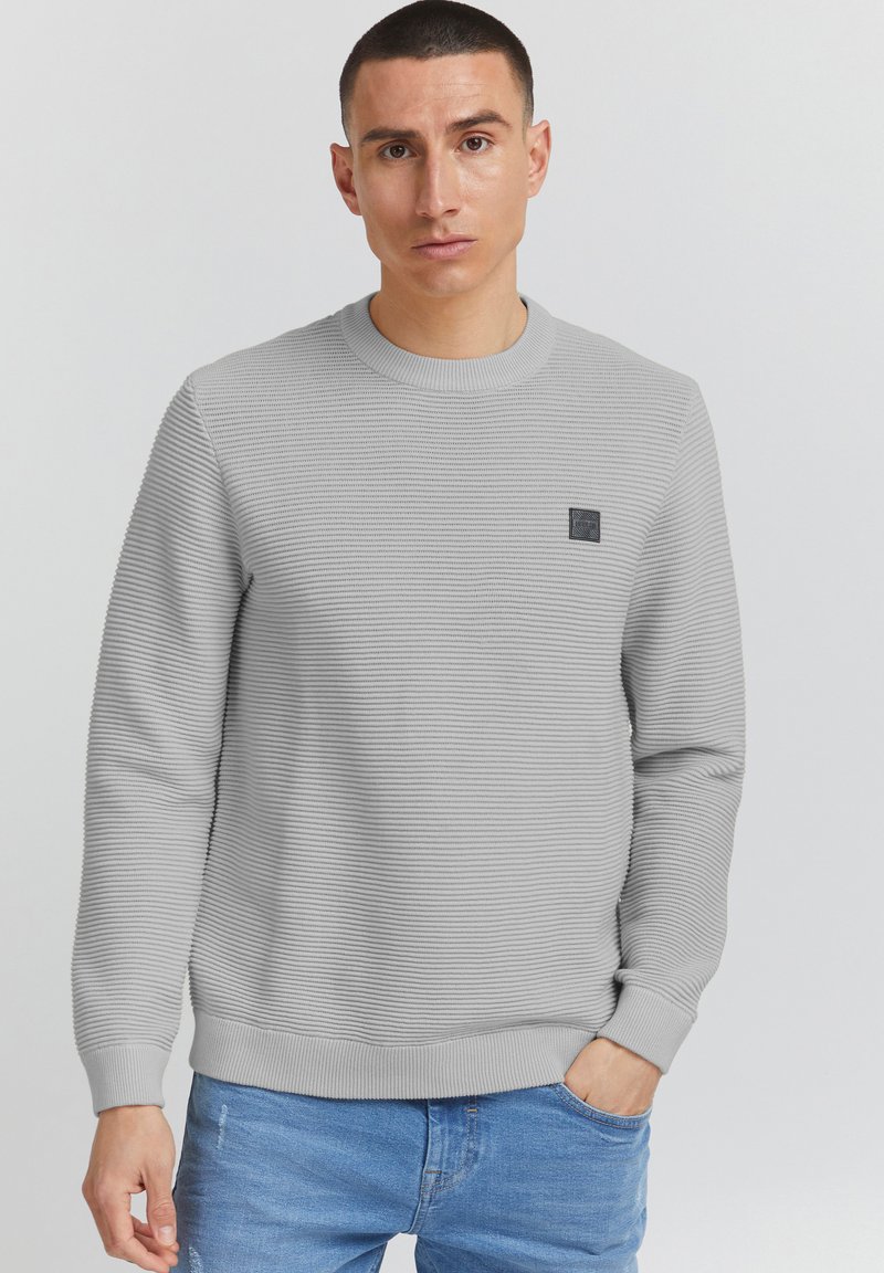 Maglione grigio chiaro con texture a coste, scollo a girocollo e una piccola etichetta nera sul petto. Abbinato a pantaloni in denim blu.