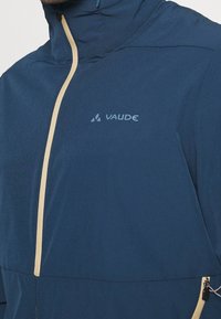 Marinblå jacka med beige dragkedjor, som har hög krage, dragkedjor på fickorna och en subtil logotyp på bröstområdet. Lättviktsmaterial.