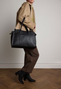 Sac en cuir noir avec deux poignées, surface texturée, forme arrondie et accents en matériel doré. Associé avec un pantalon marron et des bottes à talons noires.