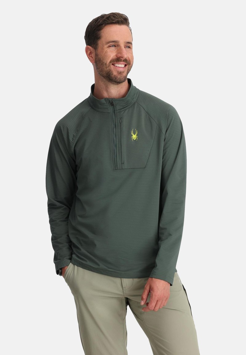 Pull vert à manches longues avec zip quart, tissu texturé, et logo araignée jaune sur la poitrine, associé à un pantalon clair.