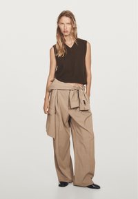 Brown sleeveless knit top paired with loose beige trousers featuring a tied waist and large pockets. Smooth texture, wide legs, and decorative buttons.