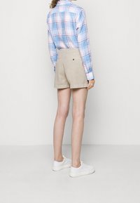 Žena v béžových šortkách, bílých teniskách a modré kostkované košili s dlouhým rukávem, stojící zády k fotoaparátu na jednobarevném pozadí.