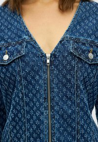 Chaqueta de mezclilla en azul oscuro con un patrón texturizado, con cuello en V, cremallera frontal y bolsillos con botón a presión con acentos de cobre.