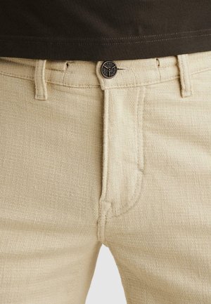 Pantalon beige texturé avec fermeture à boutons et passants pour ceinture, porté avec une chemise noire rentrée à la taille.