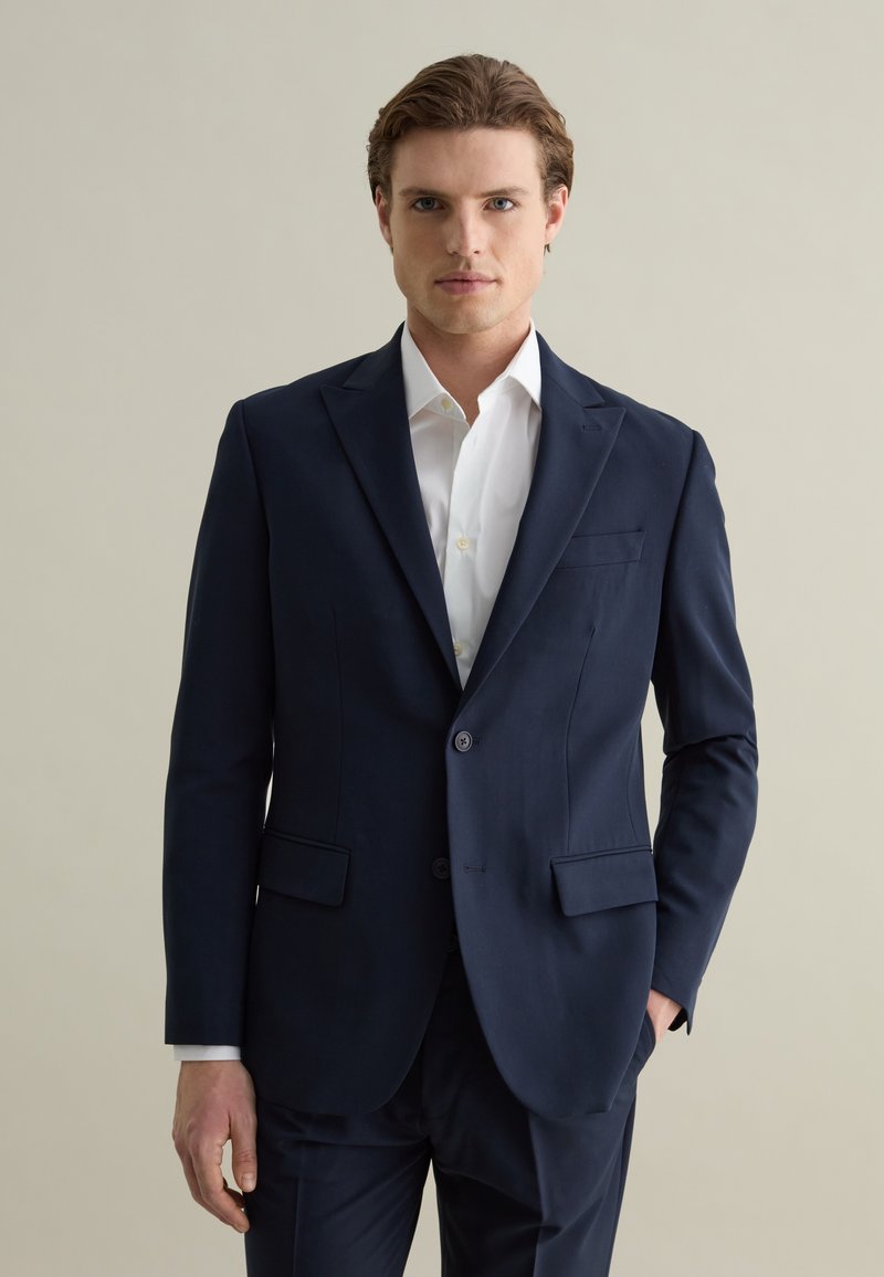 Blazer bleu marine en tissu lisse, doté de deux poches avant, d'une fermeture à bouton unique et d'un léger brillant, assorti à une chemise blanche.