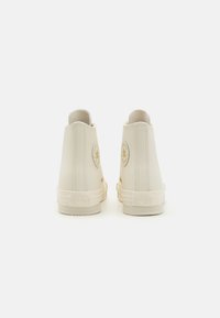 Cremfarbene High-Top-Sneaker mit glatter Textur, rundem Logo-Patch und unverwechselbarer Gummisohle mit erhabenem Branding an der Ferse.