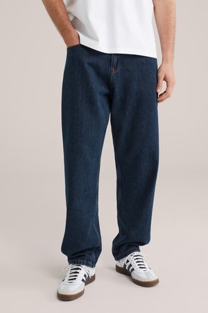 Man draagt losse donkerblauwe jeans, witte sneakers met zwarte strepen en een wit t-shirt, staat met één hand in de zak.