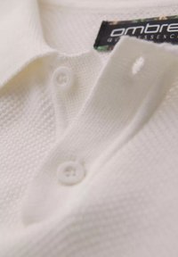 Bílý polo tričko s límečkem, který má texturovaný vzor, dva bílé knoflíky a štítek s nápisem "ombre". Detailní pohled se soustředí na detaily.
