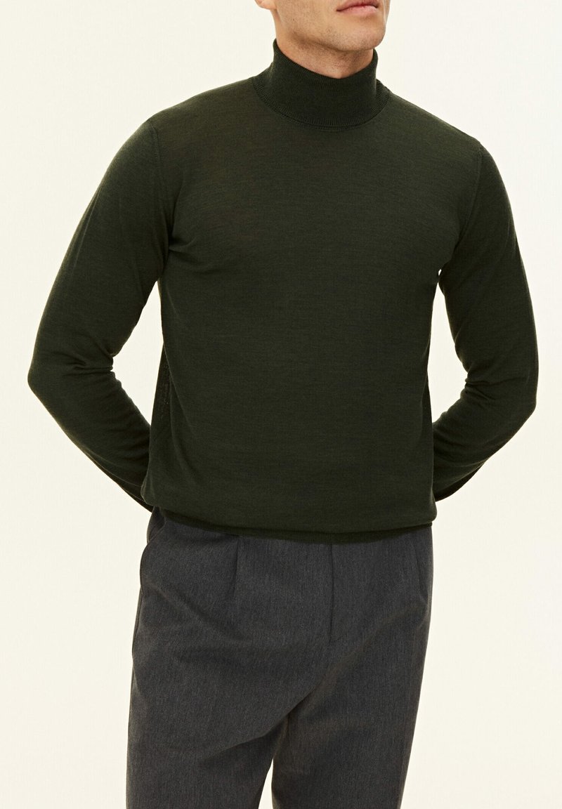 Mörkgrön turtlenecktröja, långa ärmar, åtsittande design, slät textur, i kombination med grå byxor, enkel bakgrund.