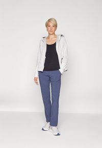 Lagani bijeli zip-up jakna s teksturiranim uzorkom, u kombinaciji s crnim majicom bez rukava i plavim hlačama; model nosi sive tenisice.