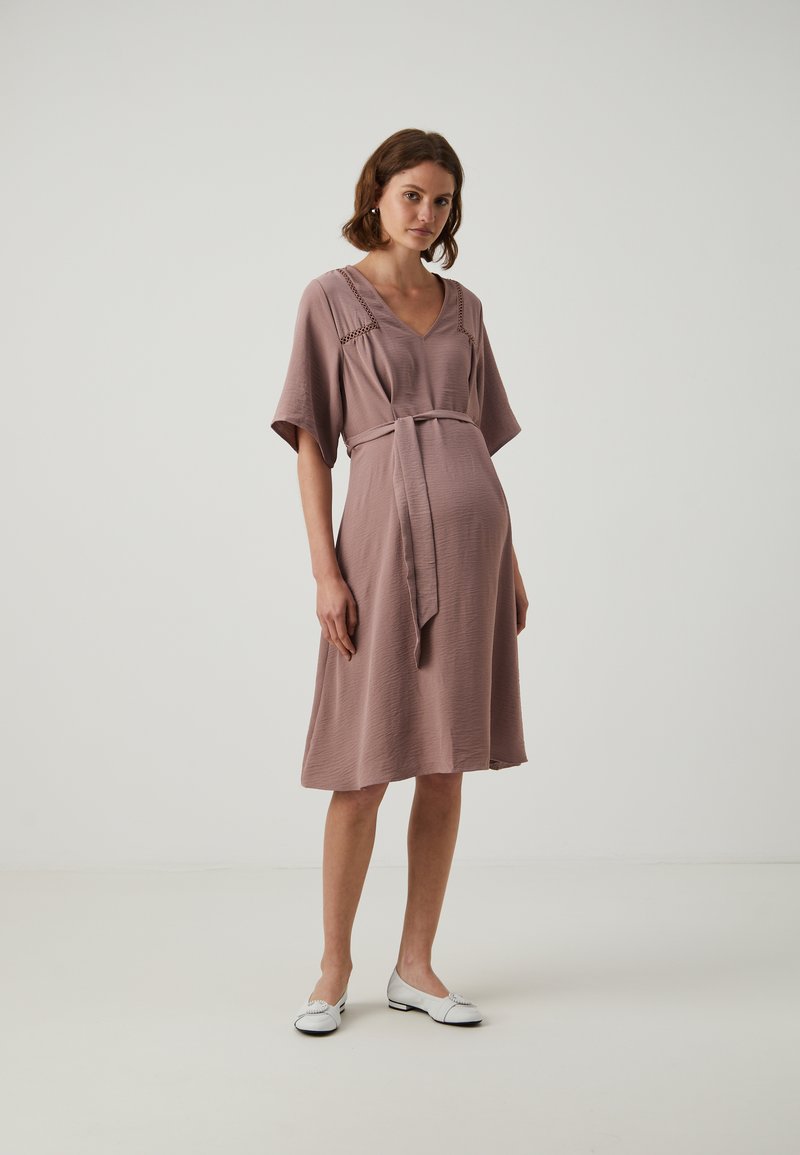 Vestido malva con escote en V y mangas cortas, que presenta un lazo en la cintura y una textura sutil. Modelo con zapatos planos blancos, de pie sobre un fondo liso.