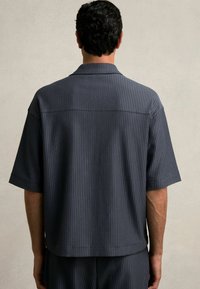 Košela s krátkym rukávom v tmavosivej farbe s vertikálnymi pruhmi. Uvoľnený strih, mäkká látka, golier a rovný lem. Minimalistický dizajn.