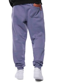 Svetle fialové tepláky s elastickými man žetami a zadní kapsou, vyrobené z měkké látky. V kombinaci s bílými sportovními botami pro ležérní vzhled.