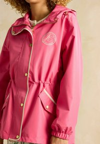 Jachetă impermeabilă roz cu manșete elastice, glugă cu șnur, buzunare cu capse și detalii cu logo. Prezintă o talie strânsă pentru a oferi formă.