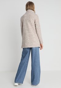 Cappotto in misto lana beige con colletto alto e finitura testurizzata, abbinato a jeans blu a gamba larga e sneakers bianche.