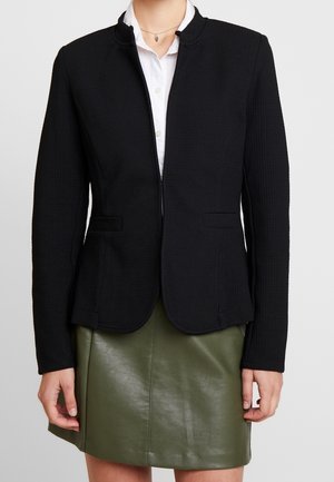 Vrouw in een zwarte getailleerde blazer over een witte shirt en een olijfgroene leren rok, staand met de armen ontspannen langs het lichaam.