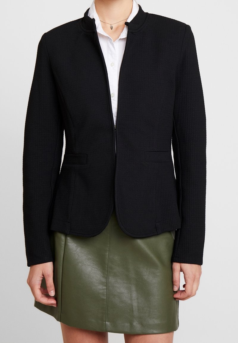 Femme portant un blazer ajusté noir sur une chemise blanche et une jupe en cuir vert olive, se tenant avec les bras détendus le long du corps.