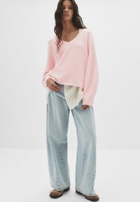 Svijetloružičasti oversized pulover s V-izrezom, složen preko bijele košulje s čipkastim detaljima, uparen s širokim svijetloplavim trapericama i smešnim cipelama.