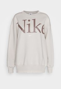 Light gray Nike sweatshirt with oversized fit, ribbed cuffs, and hem. Features large brown "Nike" logo and small white swoosh on front.