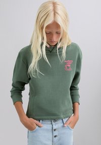 Sudadera verde con capucha y bordado rosa, bolsillo canguro y cordones. Presenta un ajuste relajado y una textura suave. Combinada con denim claro.