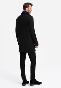 Abrigo de lana negro con cuello alto, combinado con pantalones ajustados negros y botines. Textura suave, diseño elegante y silueta larga.