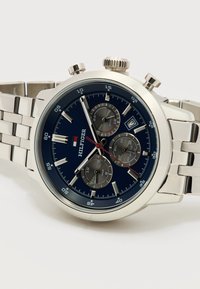 Ασημένιο μεταλλικό ρολόι χειρός με μπλε καντράν, τρεις υποκαντράν, ένδειξη ημερομηνίας και κόκκινο δείκτη δευτερολέπτων σε λευκό φόντο.