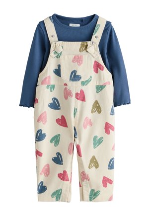 Smilende tumling med krøllet hår i to knolde, iført en blå skjorte og hvide overalls med farverige hjerter samt beige sko.
