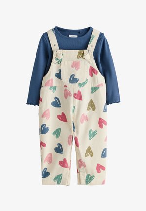 Lachende peuter met krullend haar in twee knotjes, draagt een blauw shirt en witte salopette met kleurrijke hartjes en beige schoenen.