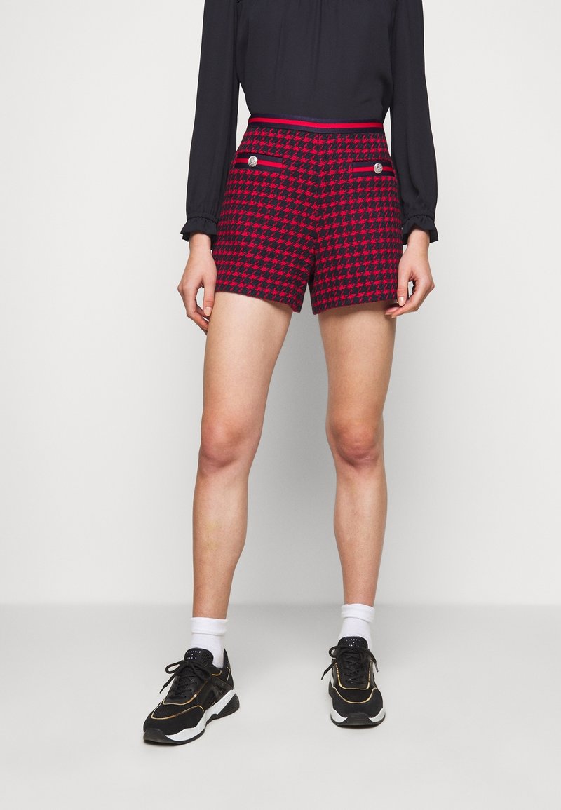 Femme portant un short rouge et noir à motif pied-de-poule avec poches à boutons, un haut à manches longues noir, des chaussettes blanches et des baskets noires.