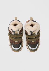 Brązowe i czarne buty za kostkę z białymi i zielonymi akcentami, posiadające miękką beżową podszewkę, rzepy oraz teksturowane gumowe podeszwy.