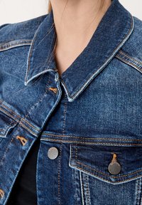 Jeansjacka i en mörkblå tvätt, med kontrasterande orange sömmar, spetsig krage och metallknappar på framfickorna.