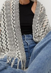 Dzianinowy szal w czarno-białe zygzakowe wzory z frędzlami na brzegach, zarzucony na czarną bluzkę i niebieskie dżinsy. Tkanina o wyraźnej fakturze.