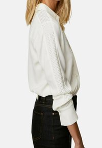 Camicia bianca a maniche lunghe con chiusura a bottoni, caratterizzata da dettagli perforati sulle spalle, abbinata a jeans di denim scuri.