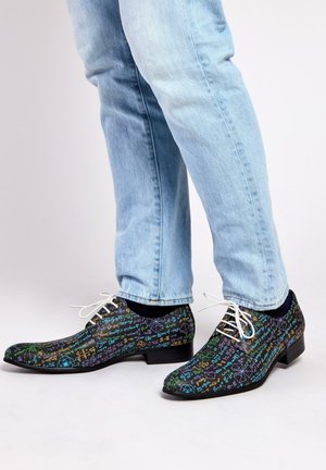 Chaussures noires habillées avec des motifs colorés de mathématiques et de sciences, à bout pointu, avec des lacets blancs et un petit talon, portées avec un jean bleu clair.