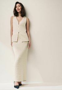 Débardeur en beige texturé avec un col en V, assorti d'une longue jupe coordonnée. La jupe a une coupe droite, accompagnée de chaussures noires à talons pointus.