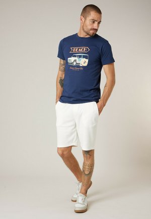 Man met tatoeages, gekleed in een marineblauw T-shirt met de tekst "Beach", witte korte broek en witte sneakers, staand met handen in de zakken, naar beneden kijkend.