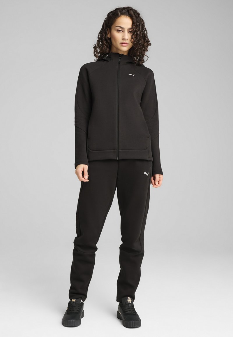 Sudadera con cremallera negra y pantalones de chándal a juego en un tejido suave y texturizado. Presenta un acento de logo, bolsillos laterales y un corte atlético. Zapatillas negras.