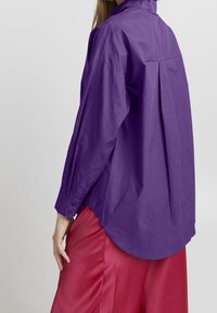 Lila Baumwollhemd mit langen Ärmeln, geknöpften Manschetten und abgerundetem Saum; verfügt über einen Rückenschlitz und wird mit einem rosa Satinrock kombiniert.