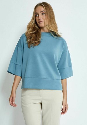Lys blå, oversized sweater med en høj hals og vide ærmer. Glat tekstur, lagdelt design, parret med beige bukser.