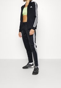 Osoba ubrana w czarną dresową kurtkę i spodnie Adidas z białymi paskami, neonowozielony biustonosz sportowy oraz czarne buty sportowe Adidas.