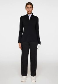 Zwart langarmig shirt met hoge kraag, ritssluiting, gecombineerd met zwarte broek en witte sportschoenen, met een golfhandschoen aan de linkerhand.