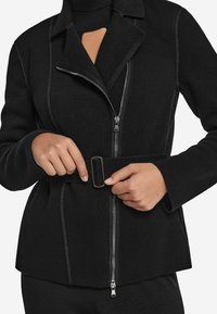 Svart jacka i mjukt tyg med dragkedja och bälte. Har en notch-krage och synliga sömnadsdetaljer.