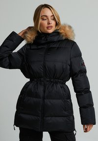 Giubbotto imbottito nero con cappuccio in pelliccia sintetica, vita elasticizzata e chiusura con zip. Presenta una texture trapuntata e un emblema sulla manica.
